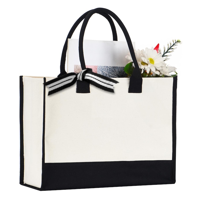 A white tote bag with a black trim, featuring a black bow tied at the top. The bag is holding a bouquet of flowers, including a white daisy and a red flower.