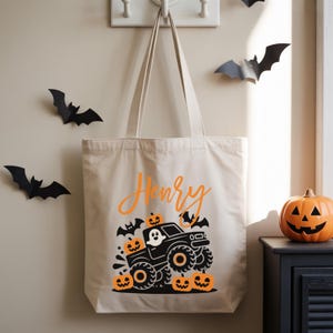 A white tote bag with a Halloween-themed design hangs on a wall, featuring a black monster truck, bats, and jack-o-lanterns.