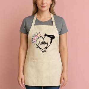 May include: A cream-coloured apron with a heart-shaped design. The design includes a floral arrangement, a hairdryer, and scissors, with the name "Ashley" in cursive. The apron has two front pockets.