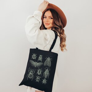 May include: A black tote bag with a white strap and a design of various insects in green on the front. The insects include a ladybug, a bee, a moth, and a beetle.