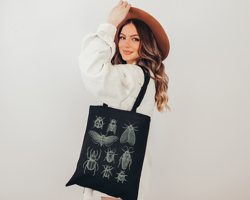 May include: A black tote bag with a white strap and a design of various insects in green on the front. The insects include a ladybug, a bee, a moth, and a beetle.