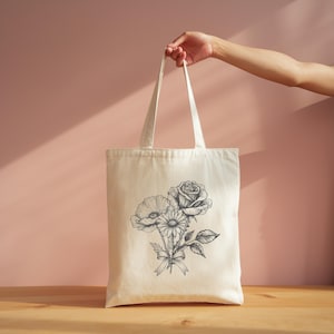 A hand holding a white tote bag with a floral design on it, against a pink background.