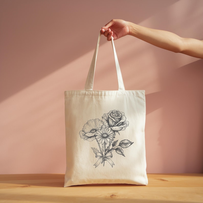 A hand holding a white tote bag with a floral design on it, against a pink background.