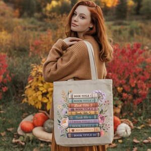 A woman with red hair stands in a field of autumn foliage, holding a tote bag with a stack of colorful books on it.