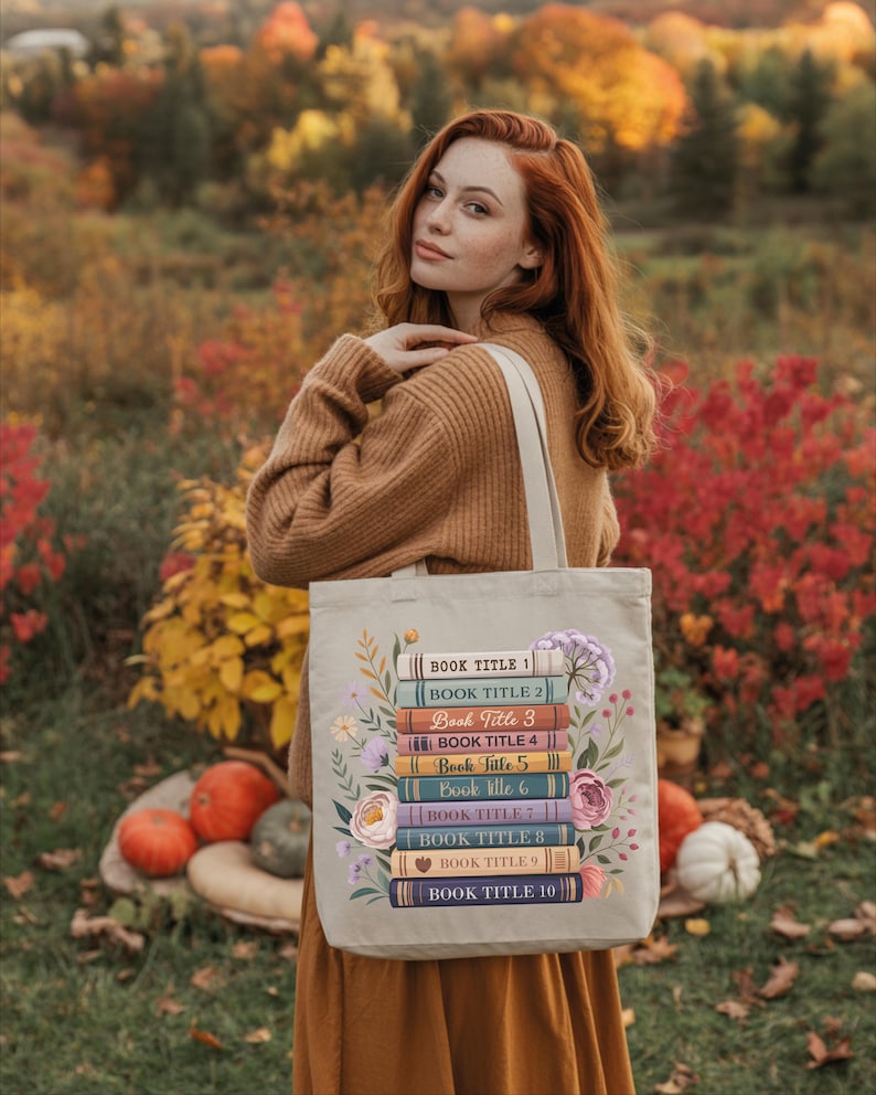 A woman with red hair stands in a field of autumn foliage, holding a tote bag with a stack of colorful books on it.