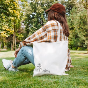 May include: A white canvas tote bag with a black rose illustration and the name "Ashley" in script. The bag has a simple design and is carried by a person wearing a brown cap and plaid shirt, sitting on green grass.