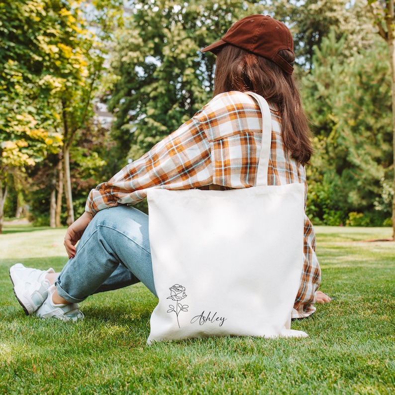 May include: A white canvas tote bag with a black rose illustration and the name "Ashley" in script. The bag has a simple design and is carried by a person wearing a brown cap and plaid shirt, sitting on green grass.