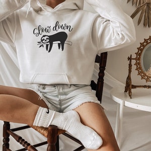 May include: White hoodie with a black sloth graphic and the text "Slow down". The model is wearing the hoodie and sitting on a wooden chair.
