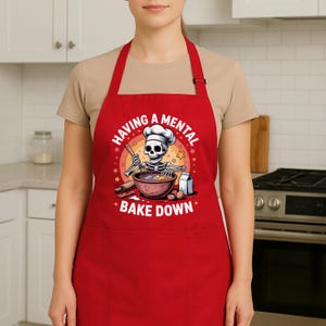 May include: A red apron featuring the phrase "HAVING A MENTAL BAKE DOWN" and a cartoon skeleton chef. The skeleton is wearing a chef's hat and mixing ingredients in a bowl. The apron has a front pocket and neck strap.