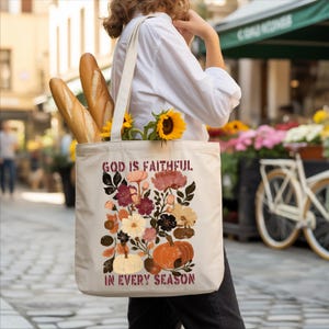 A person is walking down a street carrying a white tote bag with a floral design and text printed on it. The bag also contains a loaf of bread and a bouquet of flowers.