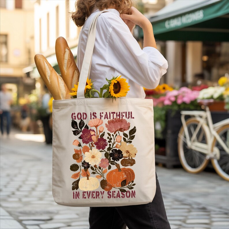 A person is walking down a street carrying a white tote bag with a floral design and text printed on it. The bag also contains a loaf of bread and a bouquet of flowers.