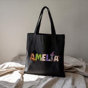 A black tote bag with the name "Amelka" printed on it, sitting on a white bedspread.