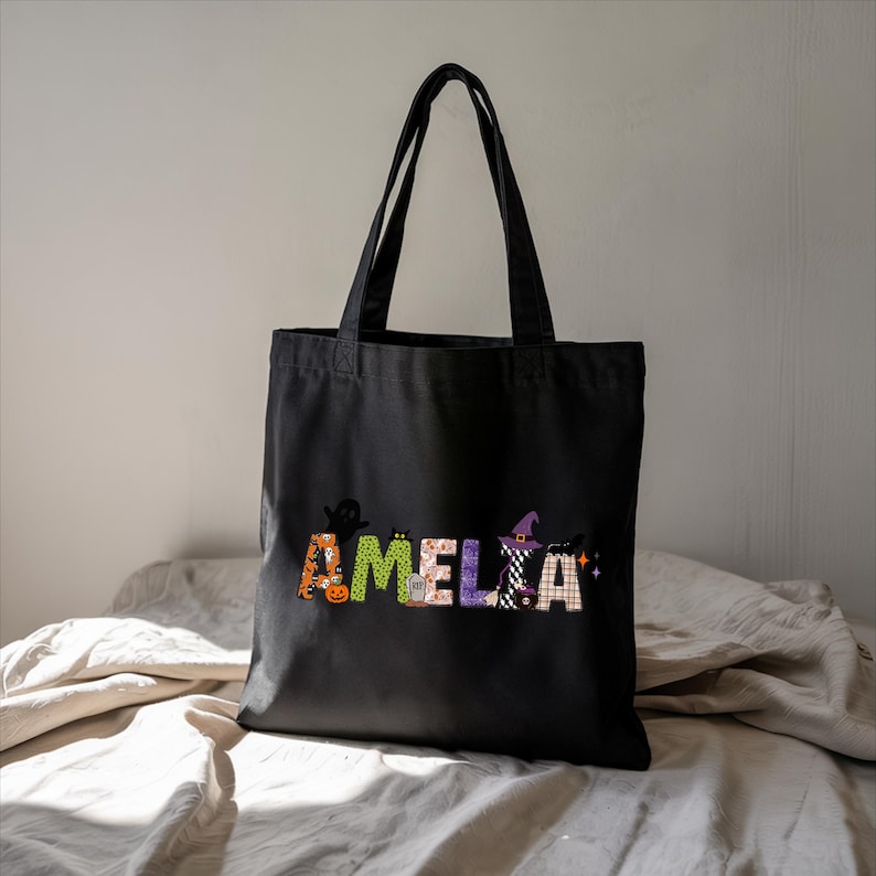 A black tote bag with the name "Amelka" printed on it, sitting on a white bedspread.