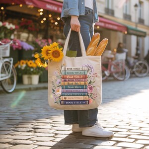 A person is standing on a cobblestone street, holding a bag decorated with a stack of colorful books and a bouquet of sunflowers.
