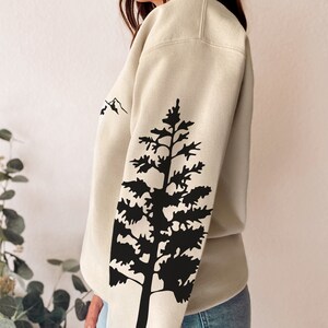 May include: A beige sweatshirt with a black silhouette of a pine tree on the sleeve. The tree is a simple design with a few branches and a trunk. The sweatshirt has a relaxed fit and a crew neck.