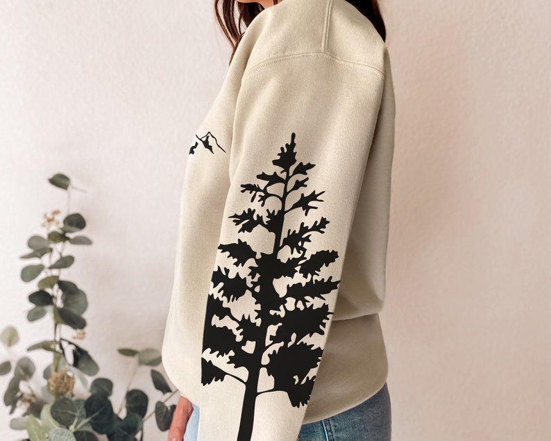 May include: A beige sweatshirt with a black silhouette of a pine tree on the sleeve. The tree is a simple design with a few branches and a trunk. The sweatshirt has a relaxed fit and a crew neck.