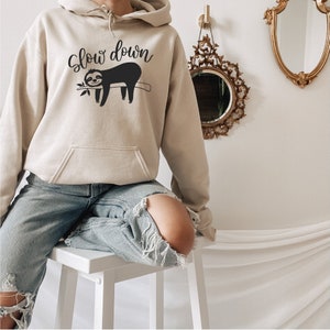 May include: A beige hoodie with a black sloth graphic and the text "Slow down". The model is wearing the hoodie with ripped jeans and is sitting on a white stool.