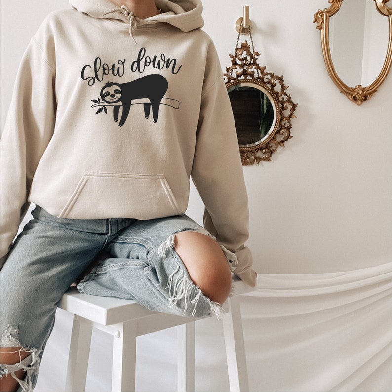 May include: A beige hoodie with a black sloth graphic and the text "Slow down". The model is wearing the hoodie with ripped jeans and is sitting on a white stool.