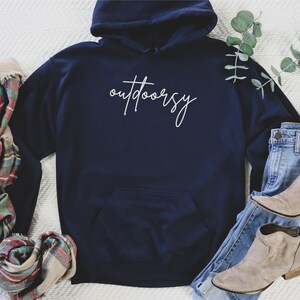 May include: A navy blue hoodie with a white cursive inscription that reads "outdoorsy". The hoodie is laid flat on a white surface with a plaid scarf, a pair of jeans, and a pair of brown ankle boots.