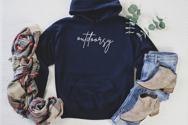May include: A navy blue hoodie with a white cursive inscription that reads "outdoorsy". The hoodie is laid flat on a white surface with a plaid scarf, a pair of jeans, and a pair of brown ankle boots.