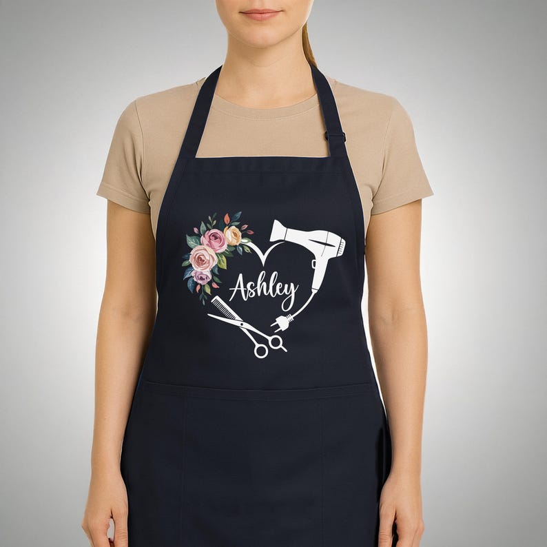 May include: A navy apron with a heart-shaped design, floral elements, and the name "Ashley". The heart includes a hairdryer, scissors, and a comb, indicating a hairstylist theme. The apron is a practical garment.