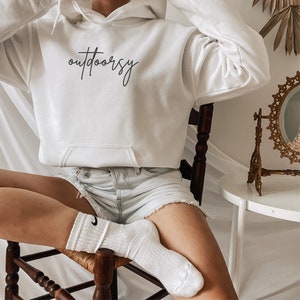 May include: A white hoodie with the word "outdoorsy" written in black cursive lettering. The hoodie is worn by a person sitting on a wooden chair.