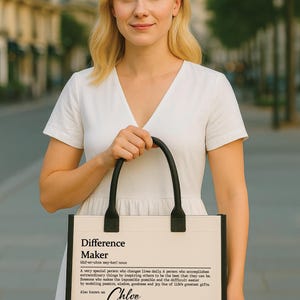 May include: A black and cream tote bag with the text "Difference Maker" and the name "Chloe" is held by a person. The bag has black handles and trim. The background is blurred, showing a city street.
