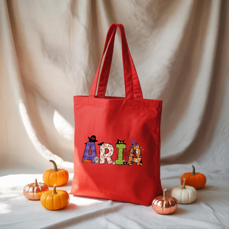 A red tote bag with the name "ARIA" printed on it, surrounded by small pumpkins.