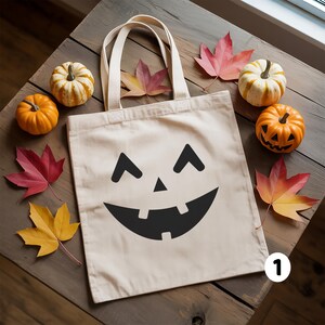 A white tote bag with a jack-o-lantern face design is placed on a wooden surface, surrounded by small pumpkins and autumn leaves.