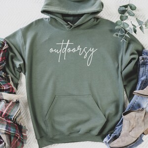 May include: A green hooded sweatshirt with the word "outdoorsy" written in white script on the front. The sweatshirt is laid flat on a white surface with a plaid scarf, a pair of jeans, and two pairs of brown ankle boots.
