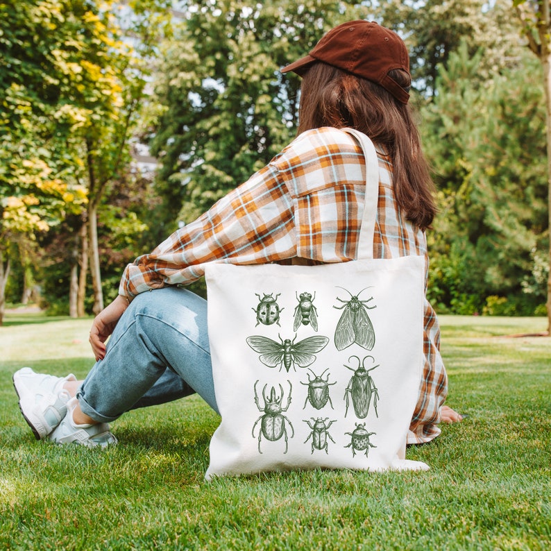 May include: A white tote bag featuring a green insect design. The bag has a long handle and is made of canvas. The insects are drawn in a vintage style and include beetles, butterflies, and moths.