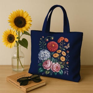 May include: A navy blue tote bag with a floral design in red, pink, yellow, and blue. The bag has two handles and is on a wooden surface next to a book and sunglasses. Two sunflowers are in a glass vase.