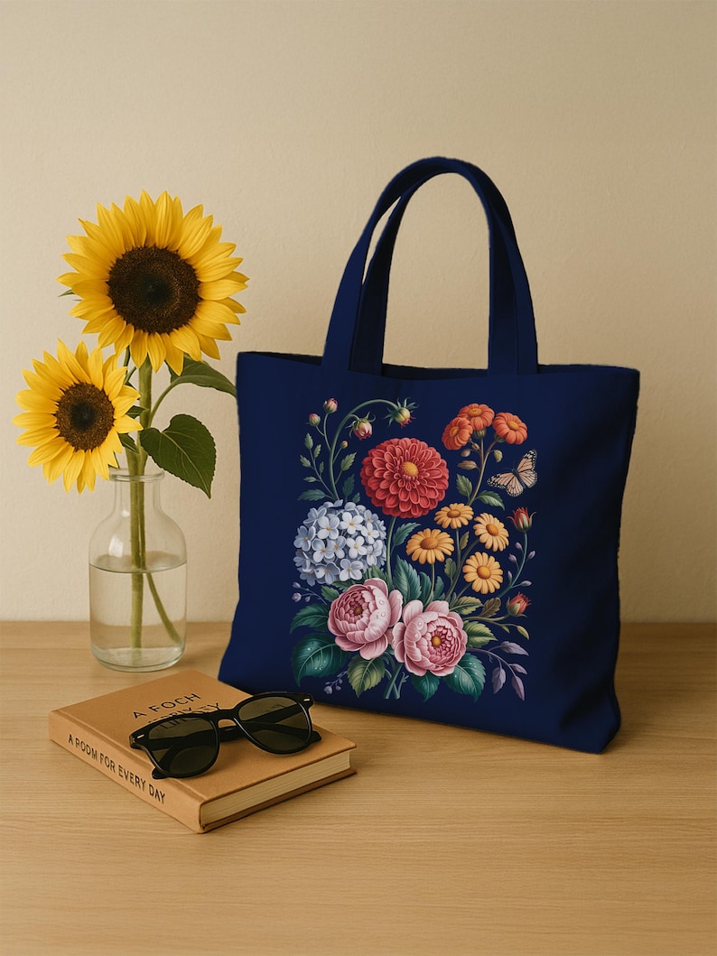 May include: A navy blue tote bag with a floral design in red, pink, yellow, and blue. The bag has two handles and is on a wooden surface next to a book and sunglasses. Two sunflowers are in a glass vase.