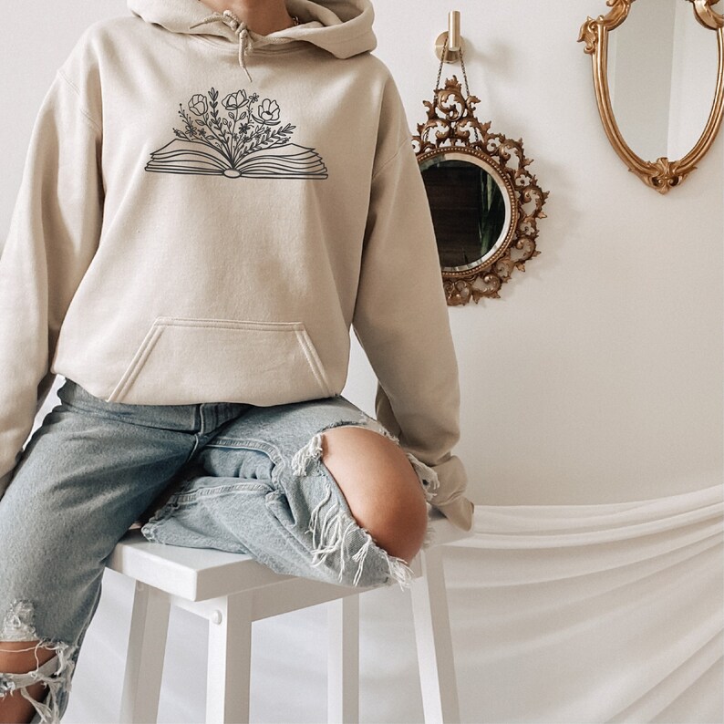 May include: A beige hoodie with a black outline of an open book with flowers growing out of it.