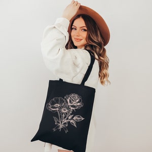 K&ouml;nnte beinhalten: Eine schwarze Tragetasche mit einem rosafarbenen Blumendesign. Die Tasche hat einen langen Riemen und wird von einer Person getragen, die einen wei&szlig;en Pullover und einen braunen Hut tr&auml;gt.