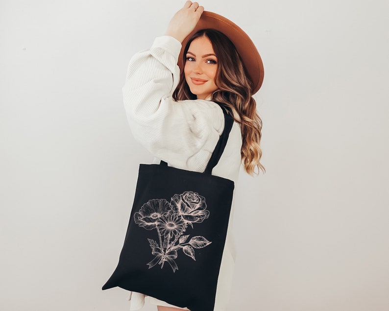 K&ouml;nnte beinhalten: Eine schwarze Tragetasche mit einem rosafarbenen Blumendesign. Die Tasche hat einen langen Riemen und wird von einer Person getragen, die einen wei&szlig;en Pullover und einen braunen Hut tr&auml;gt.