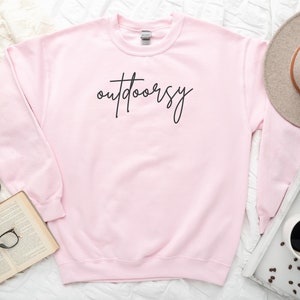 May include: A pink sweatshirt with the word "outdoorsy" written in black script on the front.