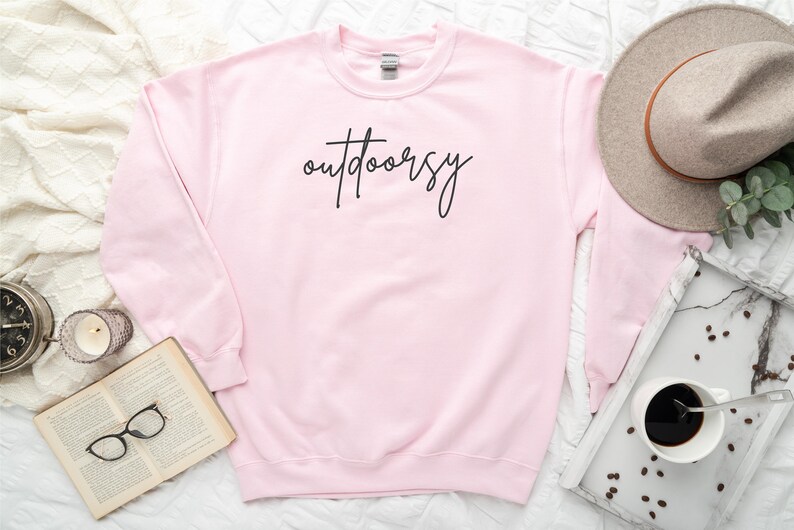 May include: A pink sweatshirt with the word "outdoorsy" written in black script on the front.