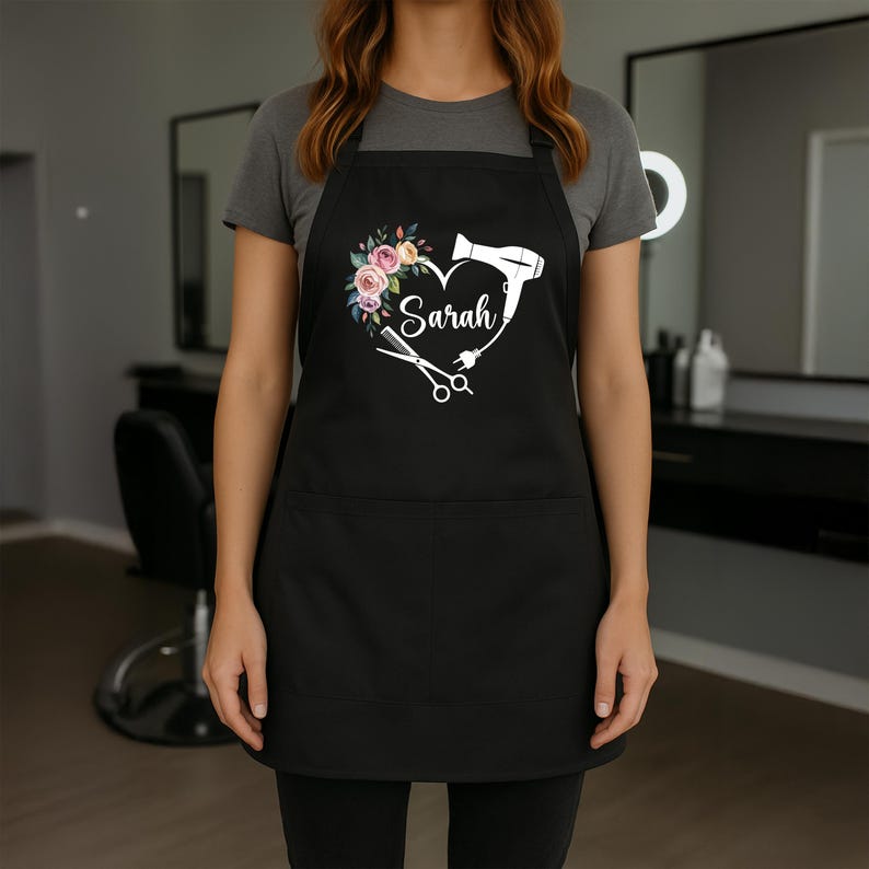 May include: Black apron with a heart-shaped design featuring a floral arrangement, a hairdryer, and scissors. The name "Sarah" is written in script within the heart. The apron has two front pockets.