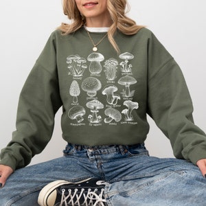 Op de afbeelding: Een groen sweatshirt met een witte grafische print van verschillende soorten paddenstoelen. De tekst op de print bevat de namen van de paddenstoelen:  porcini, parasol, honingzwam, enoki,  stuifzwam,  weboap,  moriel,  oesterzwam,  champignon,  vliegenzwam,  reishi en  zwarte trompet.