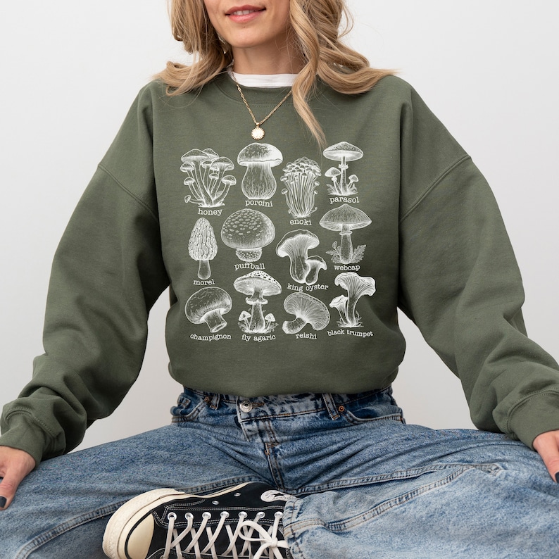 Op de afbeelding: Een groen sweatshirt met een witte grafische print van verschillende soorten paddenstoelen. De tekst op de print bevat de namen van de paddenstoelen:  porcini, parasol, honingzwam, enoki,  stuifzwam,  weboap,  moriel,  oesterzwam,  champignon,  vliegenzwam,  reishi en  zwarte trompet.