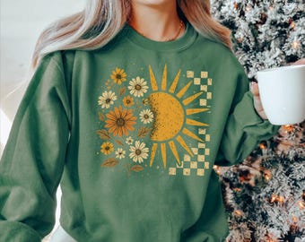 May include: Green sweatshirt featuring a floral and sun graphic. The design includes a yellow sun, daisies, and a checkered pattern. The sweatshirt is a solid green color.