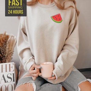 Op de afbeelding: Een lichtbeige sweatshirt met een watermeloenprint op de voorkant. Het model draagt ​​een gescheurde spijkerbroek en houdt een roze mok vast.
