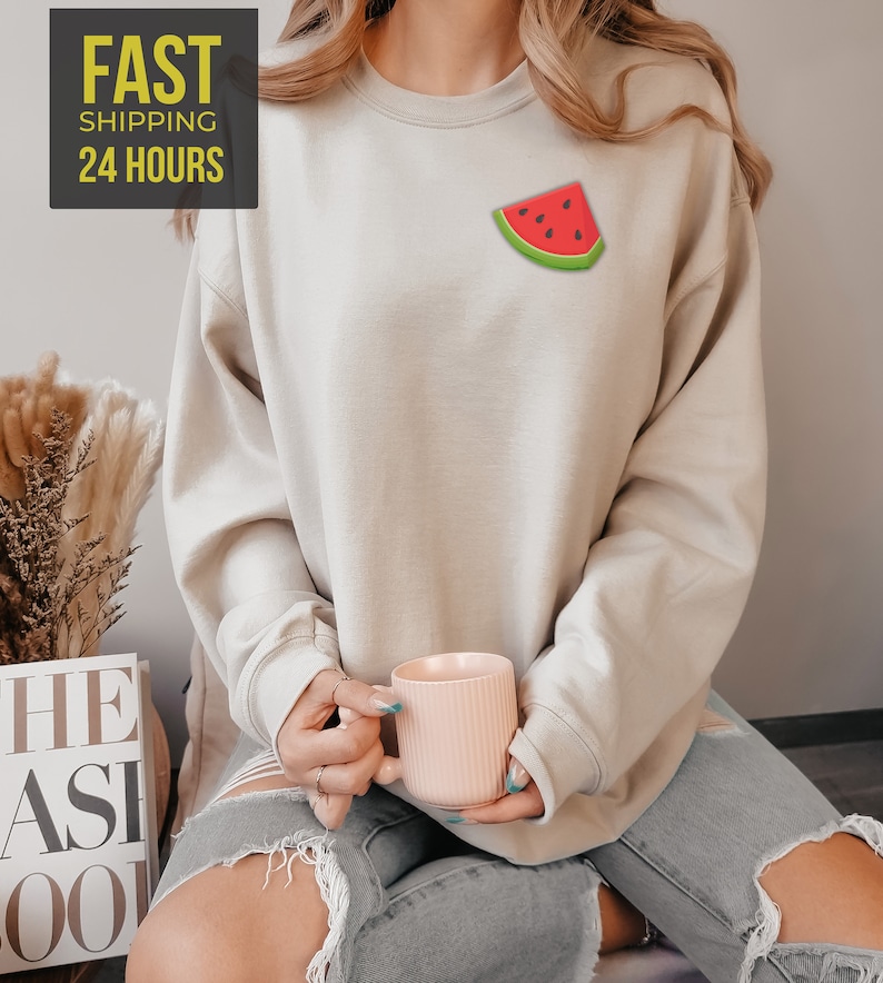 Op de afbeelding: Een lichtbeige sweatshirt met een watermeloenprint op de voorkant. Het model draagt ​​een gescheurde spijkerbroek en houdt een roze mok vast.