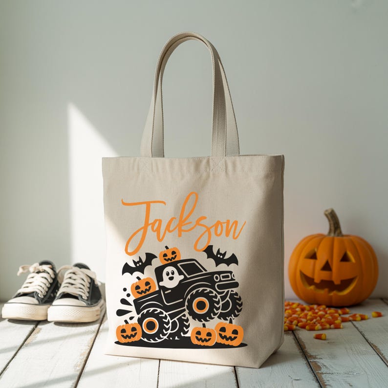 A beige tote bag with a Halloween-themed design, featuring a monster truck and jack-o-lanterns, is placed on a wooden surface. In the background, there are a pair of shoes and a carved jack-o-lantern.