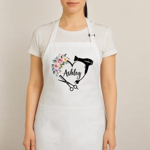 May include: White apron with a heart-shaped design incorporating a floral arrangement, a hairdryer, and scissors. The name "Ashley" is written in cursive within the heart. The apron has a neck strap and waist ties.