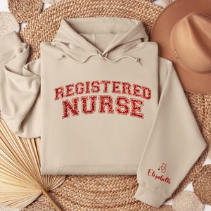 May include: Beige hoodie with the text "REGISTERED NURSE" in red glitter lettering. The sleeve has a small red stethoscope and the name "Elizabeth" in red lettering.