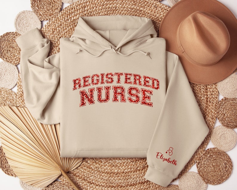 May include: Beige hoodie with the text "REGISTERED NURSE" in red glitter lettering. The sleeve has a small red stethoscope and the name "Elizabeth" in red lettering.