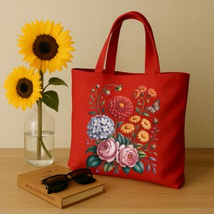May include: A red tote bag with a floral design featuring pink, blue, and yellow flowers, and a butterfly. Two sunflowers in a glass vase and a book with sunglasses rest on a wooden surface.