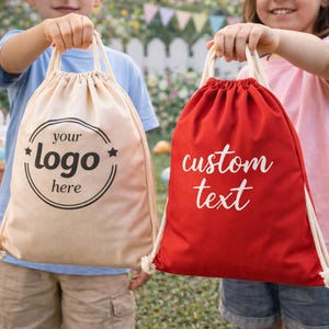 May include: Two drawstring bags are held up. One is beige with the text "your logo here" in a black circle. The other is red with the text "custom text" in white. Both bags have white drawstrings and are held by children.